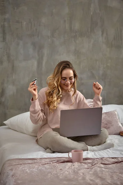 Blogger loira em óculos, roupas casuais. Sorrindo, olhando radiante, segurando cartão de plástico branco, sentado na cama com laptop nas pernas. Close-up — Fotografia de Stock