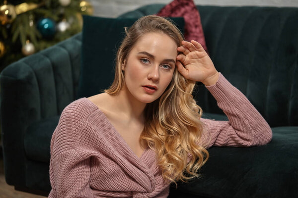 Blonde woman blogger in pink sweater. She is sitting leaning on gray sofa with colorful pillows. Interior with decorated Christmas tree. Close-up