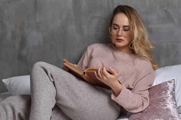 Блондинка-жінка в окулярах, повсякденний одяг лежить на ліжку, читає книгу в спальні. Студент, блогер. Інтер'єр з сірою стіною. Крупним планом — стокове фото