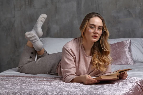 Femme blonde en lunettes, vêtements décontractés. Elle te regarde, tenant le livre, couchée sur le lit dans la chambre. Étudiant, blogueur, étudiant de documents. Gros plan — Photo