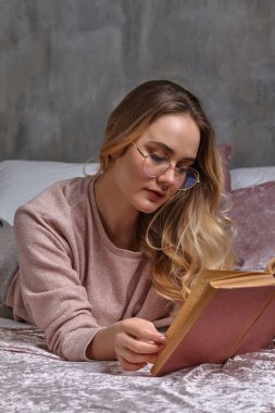 Gözlüklü, sıradan giyinen sarışın kız. O, yatakta uzanıyor ve yatak odasında kitap okuyor. Öğrenci, blogcu. İçi gri duvarla kaplı. Yakın plan.