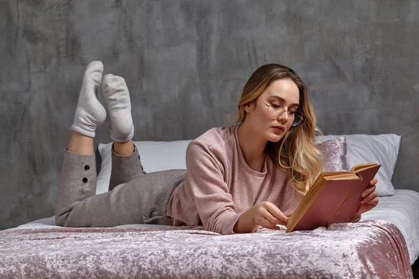 Szőke nő szemüvegben, alkalmi ruhában. Fekszik az ágyon és könyvet olvas a hálószobában. Diák, blogger. Belül szürke fallal. Közelkép — Stock Fotó