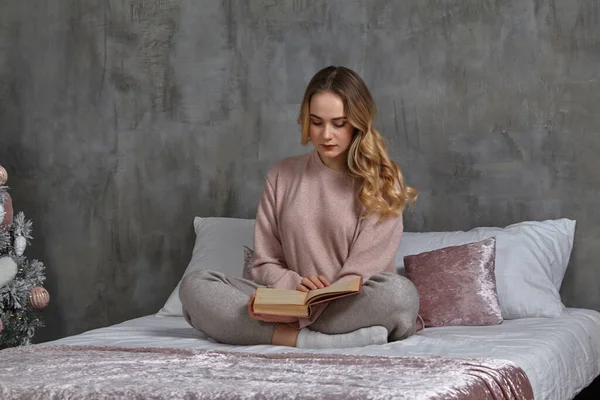Блондинка в повсякденному одязі, сидить на ліжку, читає книгу в спальні. Студент, блогер, навчальний документ. Інтер'єр з ялинкою, сіра стіна — стокове фото