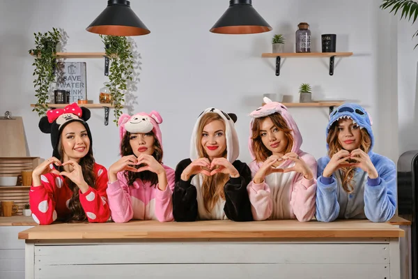 Nők öltözött plüss pizsama rajzfilmfigurák élvezi leánybúcsú, áll a konyhaasztal mögött, mutató szív jel. Közelkép. — Stock Fotó