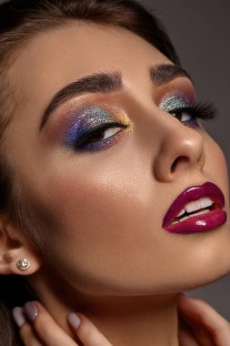 Esmer kız boynuna dokunuyor, gri arka planda yan poz veriyor. Renkli göz farı, takma kirpikler, parlak mor dudaklar ve beyaz dişler. Lüks makyaj. Profesyonel maquillage. Kapat.