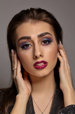 Siyah, parlak elbiseli ve takılı esmer kadın saçlarına dokunuyor, gri arka planda poz veriyor. Lüks makyaj, mükemmel cilt. Çok renkli göz farı, takma kirpikler, parlak pembe dudaklar. Kapat.