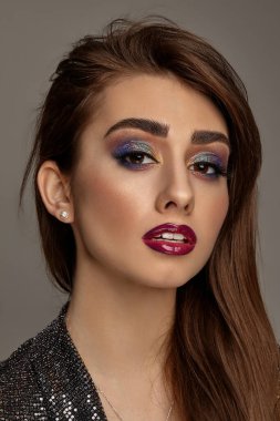 Siyah, parlak elbiseli esmer kadın sana bakıyor, gri arka planda poz veriyor. Lüks makyaj, mükemmel cilt. Renkli göz farı, takma kirpikler, parlak kırmızı dudaklar. Profesyonel maquillage. Kapat.