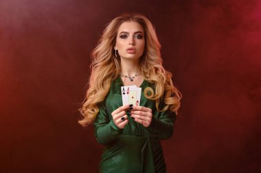 Yeşil giysili ve takılı kıvırcık sarışın kız. Renkli dumanlı arka planda poz veren iki oyun kartı gösteriliyor. Poker, kumarhane. Yakın plan.