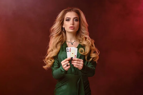 Кудрявая блондинка в зеленом стильном платье и украшениях. Показывает две игральные карты, позируя на ярком дымном фоне. Покер, казино. Крупный план — стоковое фото