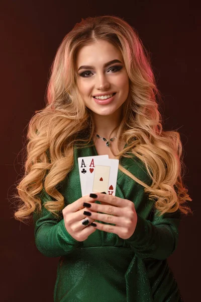 Blonďatý kudrnatý model v zelených stylových šatech a špercích. Usmívá se, ukazuje dvě esa, pózuje na hnědém pozadí. Poker, kasino. Detailní záběr — Stock fotografie