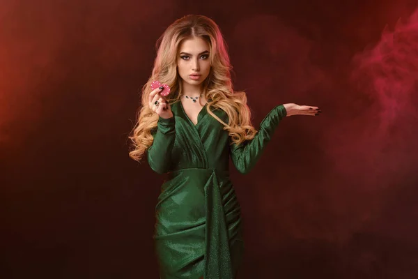 Menina loira em vestido elegante verde e jóias. Mostrando duas fichas vermelhas, segurando algo na mão, posando no fundo marrom fumado. Close-up — Fotografia de Stock