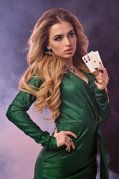 Blonďatý model v zelených šatech a špercích. Polož ruku na bok, ukaž dvě hrací karty, pózuj na fialovém zakouřeném pozadí. Poker, kasino. Detailní záběr — Stock fotografie