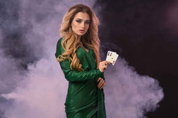 Ξανθό μοντέλο με make-up, σε πράσινο φόρεμα και κοσμήματα. Βάλτε το χέρι στο ισχίο, δείχνοντας δύο άσους, ποζάροντας σε μαύρο καπνιστό φόντο. Πόκερ, καζίνο. Κοντινό πλάνο — Φωτογραφία Αρχείου