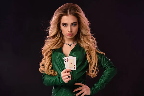 Blonde model in green stylish dress and jewelry. Put her hand on hip, showing two playing cards, posing on black background. Poker, casino. Close-up