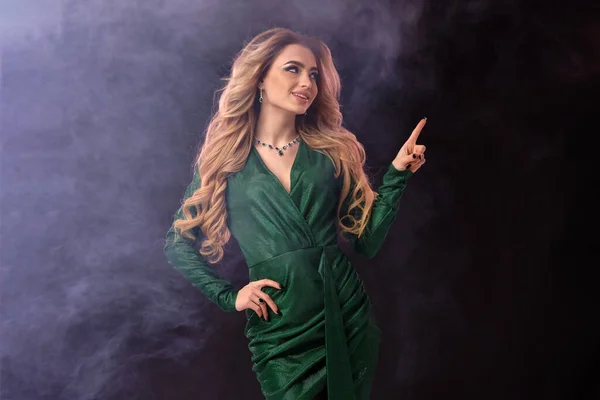 Ξανθιά γυναίκα σε πράσινο κομψό φόρεμα και κοσμήματα. Χαμογελώντας, δείχνοντας κάτι με δείκτη, ποζάροντας σε μαύρο καπνιστό φόντο. Κλείσιμο, αντιγραφή χώρου — Φωτογραφία Αρχείου
