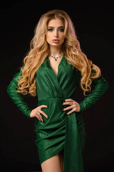 Блондинка кучерява дама в зеленій стильній сукні та ювелірних виробах. Вона поклала руки на талію, позуючи на чорному студійному фоні. Мода і краса. крупним планом — стокове фото