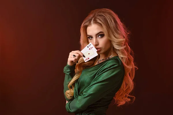 Blonde Frau in grünem stylischem Kleid und Schmuck. Zeigt zwei Spielkarten, die seitlich auf braunem Studiohintergrund posieren. Poker, Casino. Nahaufnahme — Stockfoto
