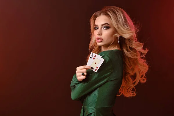 Blonde Frau in grünem stylischem Kleid und Schmuck. Sie faltete ihre Hände, zeigte zwei Asse und posierte seitlich auf rotem Studiohintergrund. Poker, Casino. Nahaufnahme — Stockfoto