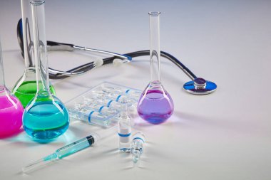 Renkli kimyasal reaksiyonlu dört tıbbi şişe. Beyaz üzerinde şeffaf sıvı, şırınga ve fonendoskop bulunan yedi model ampul. Coronavirus 'un laboratuvar araştırması, COVID-19. Kapat.