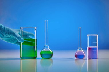 Yeşil sıvıyla tek kullanımlık eldiven taşıyın. Mor kimyasal reaksiyonlu iki tıbbi matara, mavi arka plan. Coronavirus laboratuvar araştırması. Dünya çapında salgın COVID-19. Kapat.