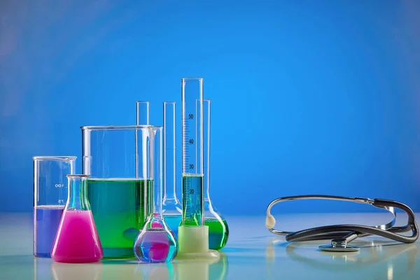 Colorful chemical reagents in beakers, medical flasks, measuring cylinder and phonendoscope, blue background. Coronavirus laboratory research. Worldwide pandemic COVID-19. Infectious disease. Close up