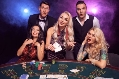 Bir grup pozitif arkadaş kumarhanede poker oynuyor. Gençler büyük bir galibiyet için bahis oynuyorlar. Siyah dumanlı arka planda kırmızı ve mavi ışıklara karşı masayı poz verirken gülümsüyorlar. Kartlar, fişler, para, kumar, eğlence ortaklığı