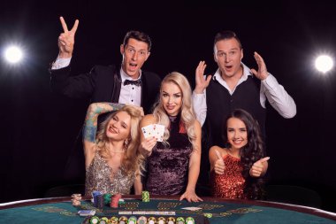 Bir grup genç ve sevimli arkadaş kumarhanede poker oynuyorlar. Gençler büyük bir galibiyet için bahis oynuyorlar. Masada siyah arka plandaki beyaz ışıklara karşı poz vermek onları heyecanlandırıyor. Kartlar, çipler, para, kumar, eğlence konsepti.