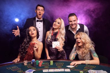 Bir grup pozitif iş arkadaşı kumarhanede poker oynuyor. Gençler büyük bir galibiyet için bahis oynuyorlar. Gülümsüyorlar ve siyah dumanlı arka planda kırmızı ve mavi arka farlara karşı masada poz vermekten memnun görünüyorlar. Kartlar, çipler, para