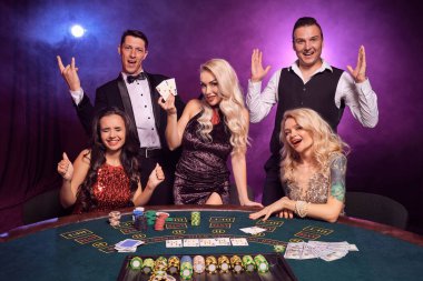 Bir grup mutlu genç kumarhanede poker oynuyor. Gençler büyük bir galibiyet için bahis oynuyorlar. Gülümsüyorlar ve siyah dumanlı arka plandaki kırmızı ve mavi ışıklara karşı masada poz veren kameraya bakıyorlar. Kartlar, çipler, para, 