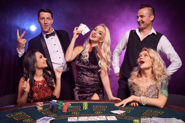Genç ve zengin bir grup kumarhanede poker oynuyor. Gençler büyük bir galibiyet için bahis oynuyorlar. Gülümsüyorlar ve siyah dumanlı arka planda kırmızı ve mavi arka farlara karşı masada poz verirken mutlu görünüyorlar. Kartlar, fişler, para, kumar, 
