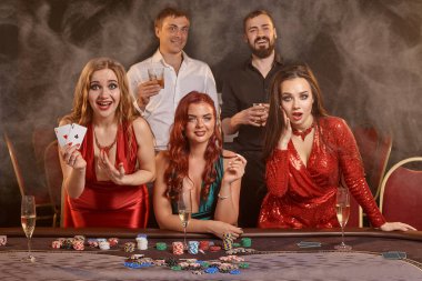 Şanslı arkadaşlar kumarhanede poker oynuyorlar. Kazanmalarını kutluyorlar, gülümsüyorlar ve karanlık bir duman geçmişine karşı masada poz veriyorlar. Kartlar, çipler, para, alkol, kumar, eğlence konsepti.