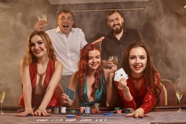 Neşeli meslektaşlar kumarhanede poker oynuyorlar. Kazanmalarını kutluyorlar, gülümsüyorlar ve karanlık bir duman geçmişine karşı masada poz veriyorlar. Kartlar, fişler, para, alkol, servet, kumar, eğlence konsepti.