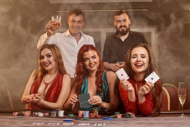 Neşeli sınıf arkadaşları kumarhanede poker oynuyorlar. Kazanmalarını kutluyorlar, gülümsüyorlar ve karanlık bir duman geçmişine karşı masada poz veriyorlar. Kartlar, fişler, para, alkol, servet, kumar, eğlence konsepti.