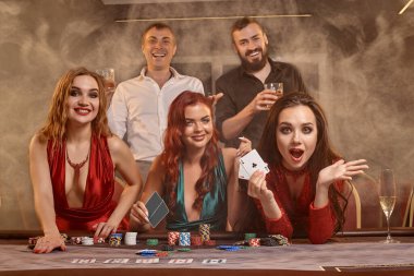 Neşeli arkadaşlar kumarhanede poker oynuyorlar. Kazanmalarını kutluyorlar, gülümsüyorlar ve karanlık bir duman geçmişine karşı masada poz veriyorlar. Kartlar, fişler, para, alkol, servet, kumar, eğlence konsepti.