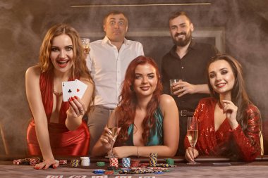 Mutlu sınıf arkadaşları kumarhanede poker oynuyorlar. Kazanmalarını kutluyorlar, gülümsüyorlar ve karanlık bir duman geçmişine karşı masada poz veriyorlar. Kartlar, fişler, para, alkol, servet, kumar, eğlence konsepti.