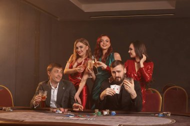 Neşeli arkadaşlar kumarhanede poker oynuyorlar. Kazanmalarını kutluyorlar, gülümsüyorlar ve karanlık bir duman geçmişine karşı masada poz veriyorlar. Kartlar, fişler, para, alkol, servet, kumar, eğlence konsepti.