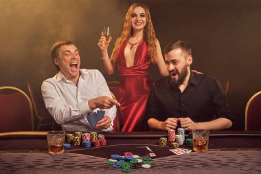 İki yakışıklı arkadaş ve muhteşem bayan kumarhanede poker oynuyorlar. Kazandıkları için mutlular, seviniyorlar ve gülümsüyorlar. Masada arka plandaki sarı ışığa karşı poz veriyorlar. Kartlar, çipler, para, kumar, eğlence konsepti.