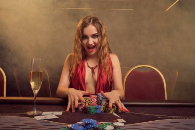 Uzun kıvırcık saçlı, mükemmel makyajlı, seksi kırmızı elbiseli çekici bir kadın. Kumar masasında oturuyor, kumar fişleri tutuyor ve gülümsüyor. Poker konsepti, sarı bir spot ışığı altında karanlık bir duman arka planında. Kumarhane.