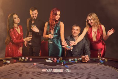 Mutlu arkadaşlar kumarhanede poker oynuyorlar. Zaferlerini kutluyorlar, gülümsüyorlar ve çok heyecanlı görünüyorlar. Bir sahne ışığı altında, karanlık bir duman arka planına karşı poz veriyorlar. Kartlar, çipler, para, alkol, kumar, eğlence konsepti