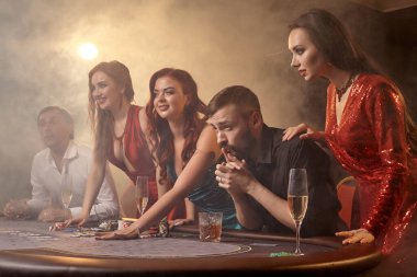 Neşeli yoldaşlar kumarhanede poker oynuyorlar. Gençler büyük bir galibiyet için iddiaya girerken masada siyah bir dumanla beyaz bir spot ışığına karşı poz veriyorlar. Kartlar, çipler, para, alkol, kumar, eğlence konsepti.