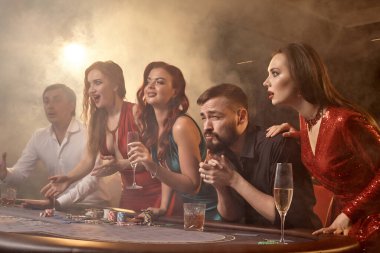 Neşeli arkadaşlar kumarhanede poker oynuyorlar. Gençler büyük bir galibiyet için iddiaya girerken masada siyah bir dumanla beyaz bir spot ışığına karşı poz veriyorlar. Kartlar, çipler, para, alkol, kumar, eğlence konsepti.
