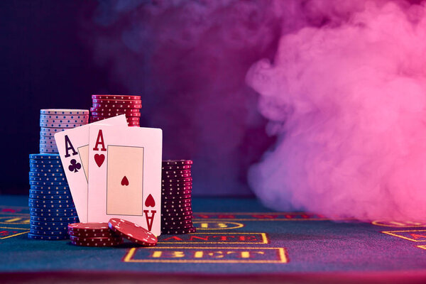 Two aces, hearts and clubs, are standing leaning on multicolored chips piles on a blue cover of a playing table with markup. Black, smoke background with red and blue backlights. Gambling entertainment, playing cards, poker, casino concept. Close-up 