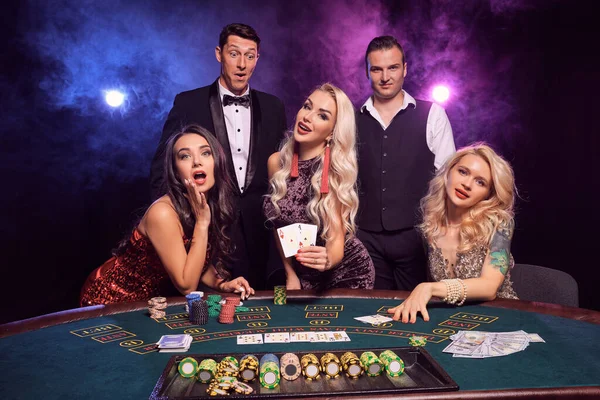 Group of a young good-looking companions are playing poker at casino. Youth are making bets waiting for a big win. They are looking satisfied posing at the table against a red and blue backlights on black smoke background. Cards, chips, money, gambli