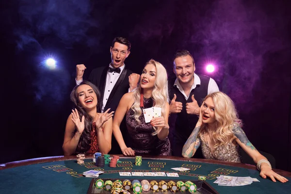 若い面白い仲間のグループは カジノでポーカーをしています 若者は大きな勝利を待って賭けをしています 黒煙を背景に赤と青のバックライトにテーブルにポーズをとって笑っている カード チップ ギャンブル 芸能人 — ストック写真