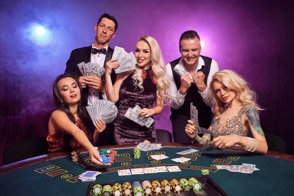 若い裕福な仲間のグループは カジノでポーカーをしています 若者は大きな勝利を待って賭けをしています 黒煙を背景に赤と青のバックライトにテーブルの上でポーズをとって笑っている カード チップ ギャンブル エンターテイメント — ストック写真