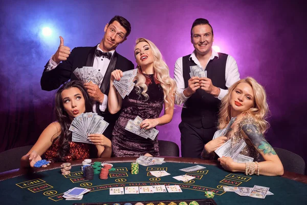 Group of a young wealthy classmates are playing poker at casino. Youth are making bets waiting for a big win. They are smiling posing at the table against a red and blue backlights on black smoke background. Cards, chips, money, gambling, entertainme