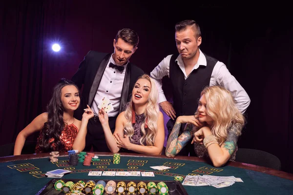 若い裕福な同僚のグループがカジノでポーカーをしています 若者は大きな勝利を待って賭けをしています 彼らは黒い背景の白いバックライトに対してテーブルでポーズをとって興奮しているように見えます カード チップ ギャンブル エンターテイメントコンス — ストック写真