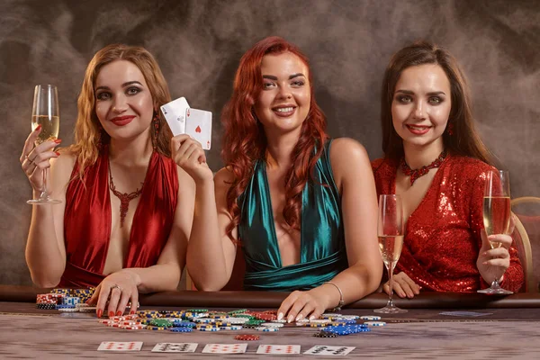 豪華な女性がカジノでポーカーをしています 彼らは彼らの勝利を祝っています カメラを見て 暗い煙の背景に対してテーブルにポーズ カード チップ アルコール ギャンブル エンターテイメントの概念 — ストック写真