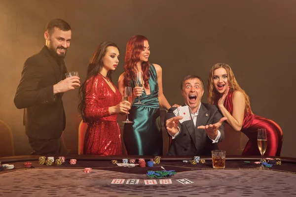 熱狂的なパートナーはカジノでポーカーをプレイしています 彼らは 暗闇の煙の背景にスポットライトを当てながらテーブルにポーズをとりながら 彼らの勝利を祝い 笑顔で 非常に興奮しています カード チップ アルコール ギャンブル — ストック写真