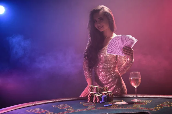 明るい化粧をしたセクシーなブルネットの女性の肖像画は 彼女の手の中に100ドル札のファンとギャンブルテーブルに対してポーズをとって 金色の輝くドレスを着て笑顔 ピンクと青と黒の煙の背景にポーカーのコンセプト — ストック写真